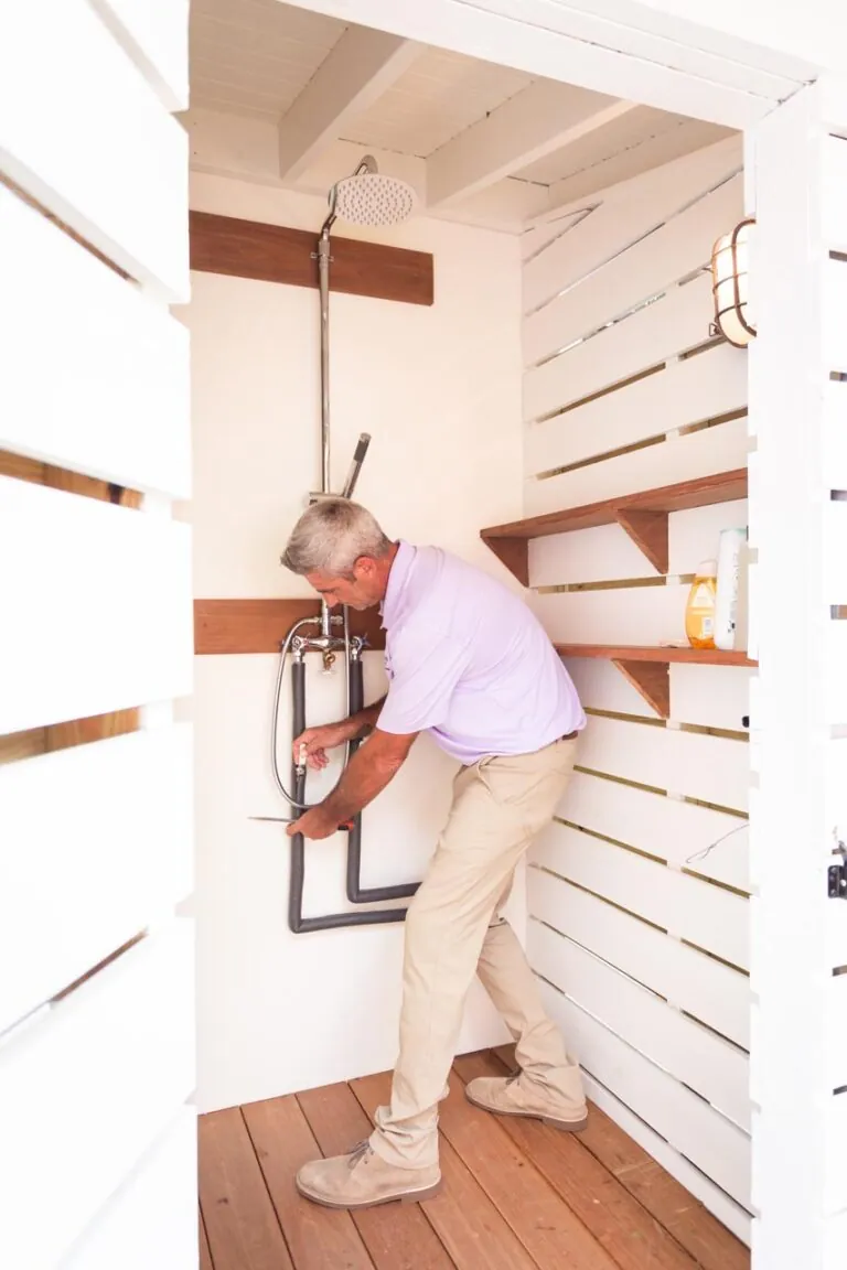 Man installing outdoor shower plumbing in white wooden enclosure. Perfect for DIY home improvement projects.