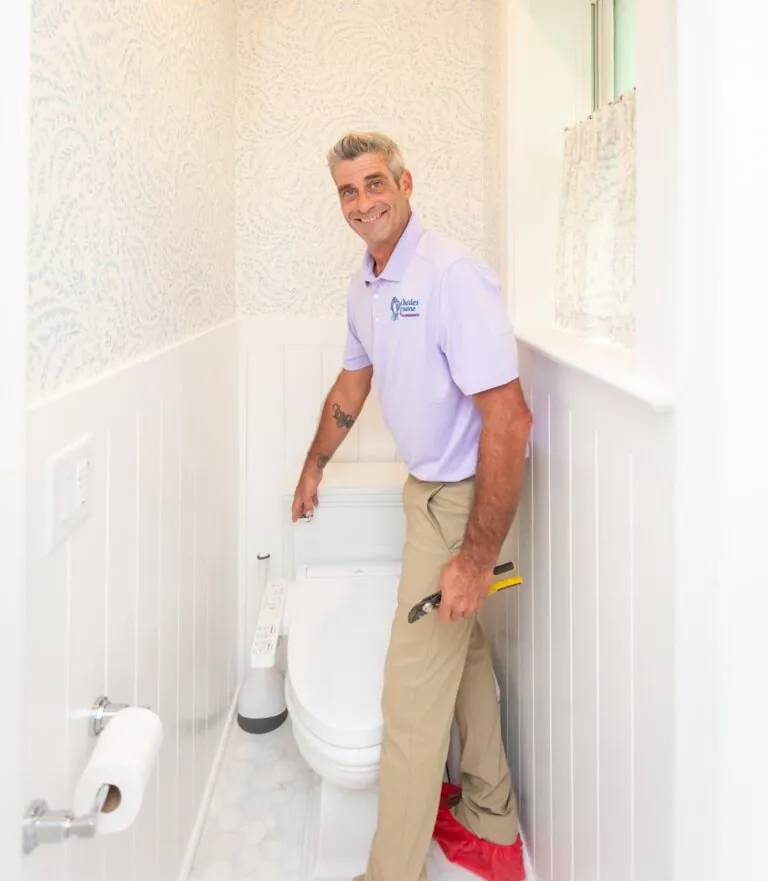 Smiling plumber installing a toilet in a bright bathroom, holding a wrench and wearing a light purple shirt.