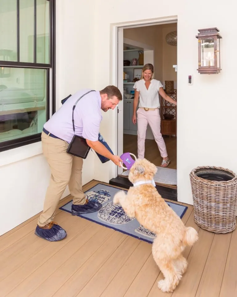 Man delivering package to woman at door while playful dog jumps excitedly.