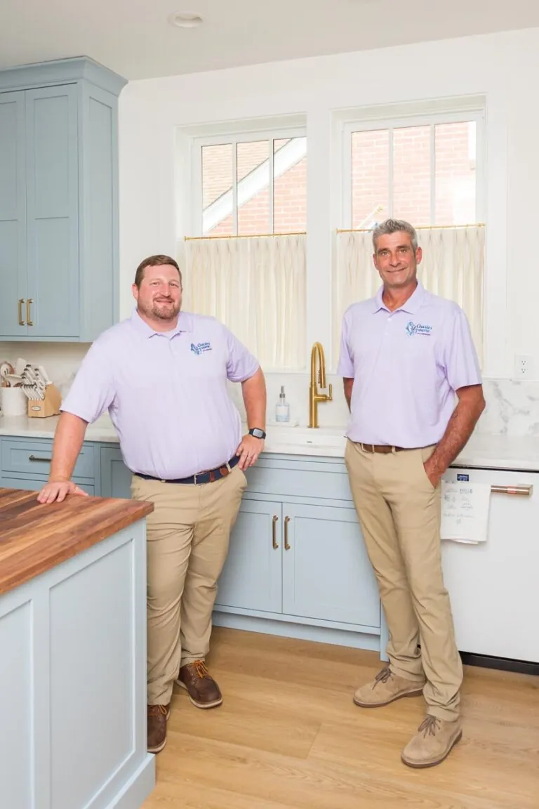 Two men in purple shirts stand in a modern kitchen with light blue cabinets and wooden countertops.