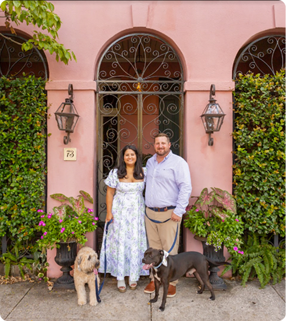 Couple with two dogs standing in front of a pink arched doorway surrounded by greenery and potted plants.